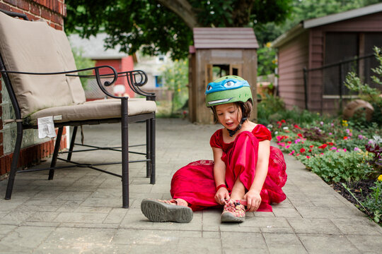 A little girl in a fancy red dress and bike helmet puts on sandal