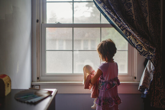 A small child stands by bedroom window tenderly holding stuffed bunny