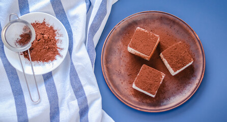 Still life with square souffle bird milk sprinkled with cocoa on a blue background top view