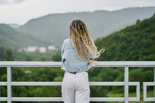 back side of a young woman with blonde braided hair wearing a denim jacket and white jean resting on a dam on a rainy day