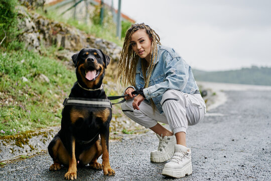 Young woman with blonde braided hair wearing a denim jacket and white jean posing next to her dog on a rainy day