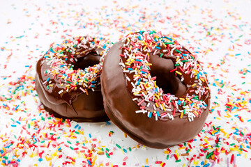 Closeup of fun chocolate donuts covered with many colored sprinkles on a white table full of sweet sprinkles