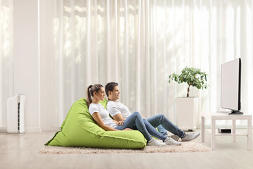 Man and woman sitting on green bean bag armchairs and watching tv at home