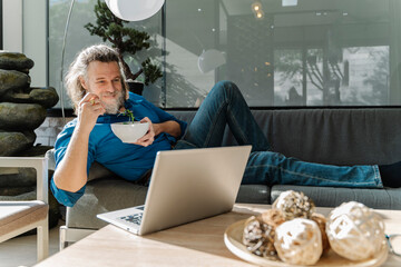 Mature man with a beard eating a salad and looking at his laptop on a