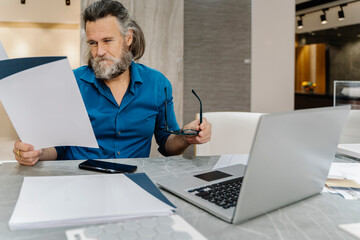 Bearded mature man reviewing some documents at his desk in front of his laptop at home. Business concept