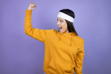 Young latin woman wearing sportswear over purple background showing arms muscles smiling proud