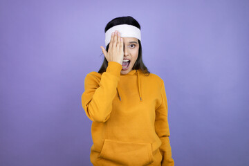 Young latin woman wearing sportswear over purple background covering one eye with hand, confident...