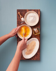womans hands holding a fork to beat eggs with various cooking ingredients on a wooden board placed on a rectangle dark wooden board on a bluish background. View from above