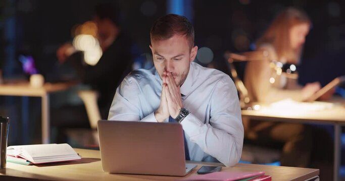 Caucasian worried young worker sitting in coworking office workspace working long hourse spending much time finishing computer project. Job stress.