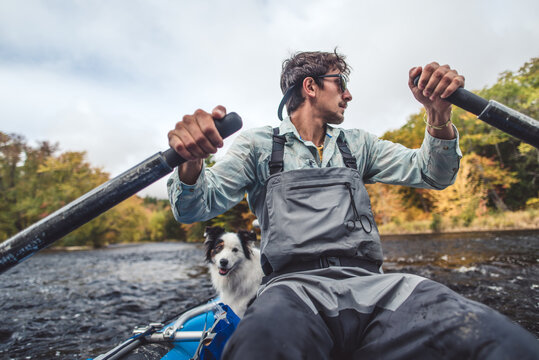 Fly fisherman rowing boat down river in fall foliage season with dog