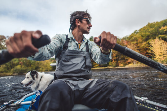 Fly fisherman rowing boat down river in fall foliage season with dog