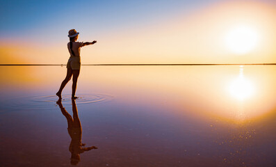 Fototapeta premium Lovely happy young girl walks and poses in a magical pose along the mirror pink salt lake enjoying the warm evening sun, looking at the fiery sunset and her reflection