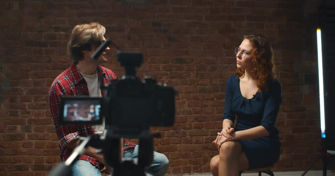 Young man entrepreneur giving interview to female journalist, camera on tripod on foreground