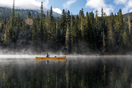 Bearded man paddles canoe on calm lake with mist rising off with trees