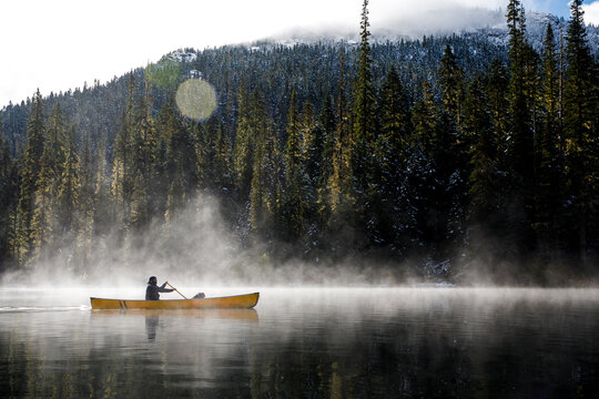 Man paddles canoe on lake by mountains with mist and fog on sunny day