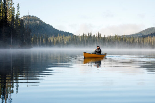 Rugged man paddles canoe on misty calm lake by thick forest