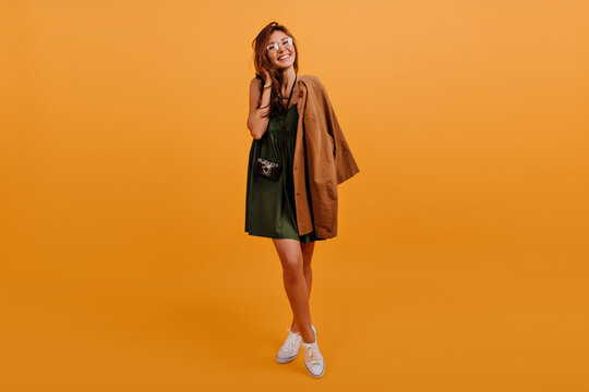 Cute shy girl with long brown hair smiles modestly and looks into camera. Full-length portrait of stylish blogger in white sneakers and dark green outfit
