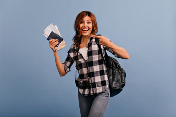 Tourist with backpack enthusiastically shows armful of tickets. Full-length shot of girl with retro camera on blue background