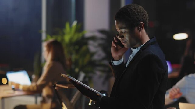 Afro-american nice professional young employee calling mobile phone servicing customer online using tablet in conference room night office.