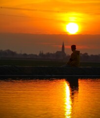 a amazing photo of a teenager sitting along the water, while the sun is setting. surrounded by farmersland, and a small town