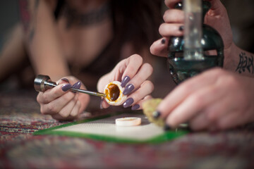 Two young ladies use a metal dabber to scoop concentrated THC cannabis wax, shatter, oil out of...