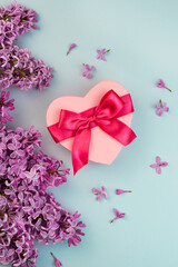 Heart shape pink gift box with red ribbon bow, purple Lilac branch flower blooming bouquet on blue background. Summer Valentine's day love creative composition. Beautiful greenery blossom