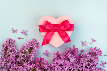 Pnk Heart shape gift box with red ribbon bow, purple Lilac branch flower blooming bouquet on blue background. Summer Valentine's day love creative composition. Blossom banner template