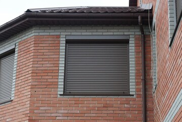 one square window is closed with brown roller shutters on the red brick wall of the attic of a...