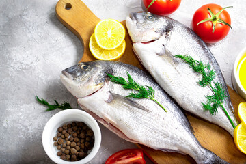 Dorado fish raw on a wooden board with cooking ingredients such as tomatoes, lemons, herbs, pepper and oil