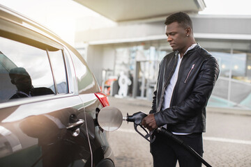 Portrait of young handsome bearded African man, wearing stylish casual clothes, refueling his luxury car at the gas station outdoors. Refueling of the car concept