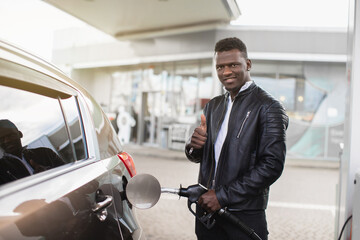 Young smiling African man refuelling his modern car at a petrol station, posing to camera with his thumb up