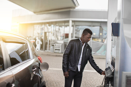 Gas petrol station. Outdoor shot of young handsome African American man in black leather jacket, refueling luxury car with gasoline, taking filling gun at the station