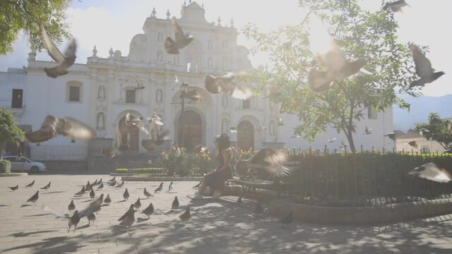Young Hispanic woman sitting in the central park of Antigua Guatemala while pigeons fly in the middle of the plaza - Woman in colonial city looking towards the cathedral 