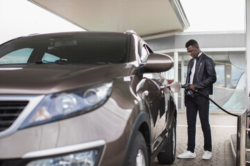 Portrait of young African American guy at outdoor gas station refueling his luxury car with gasoline, inserting nozzle in fuel cap of a car