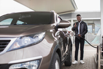 Happy bearded African man gesturing thumb up and smiling while refueling his modern luxury car on...