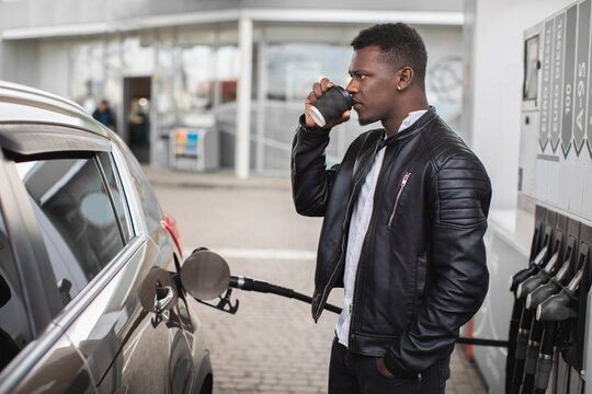 Side view of young African man, standing outdoors at the gas station, drinking take away coffee while refueling his luxury car's tank by petrol pump nozzle