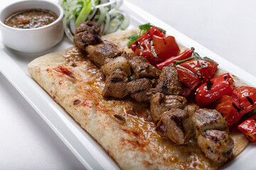 Meat shish kebab on pita bread with marinated onion sauce and baked bell pepper on a white plate on a white plate