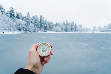 First-person view of a female hand with a compass on a background of a winter forest and a frozen lake. The concept of navigating the search for your own path and orientation to the cardinal points