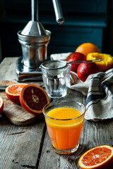 Glass of fresh blood orange juice, orange halves and juicer machine on wooden table with chair on...