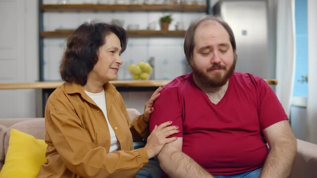 Senior woman comforting upset overweight adult son sitting together on couch