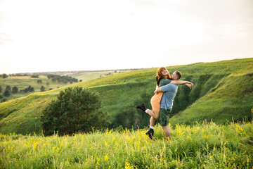 Naklejka na ściany i meble young beautiful couple red-haired girl in a pink dress and green jacket a man in a gray t-shirt and green shorts are having fun in the grass in a field in nature at sunset