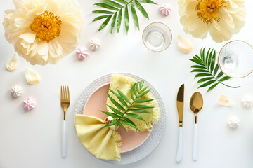 Naklejka na ściany i meble Effortless golden birthday dinner table decor. Pale yellow peony flowers and Late Spring, Summer flat lay. White dinner table, white and gold utensils, decorated with flowers, exotic palm leaves.