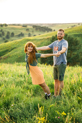 young beautiful couple red-haired girl in a pink dress and green jacket a man in a gray t-shirt and green shorts are having fun in the grass in a field in nature at sunset