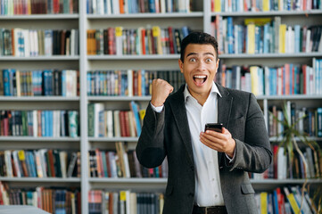 Excited hispanic male manager standing in office, uses smartphone, smiling. Happy male freelancer...
