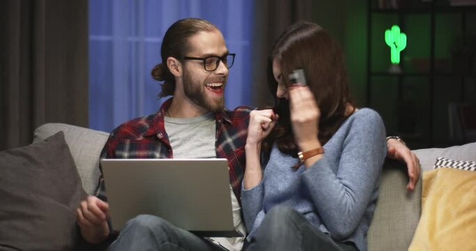 Portrait of Caucasian young cheerful happy couple wife and husband resting on couch together in living room in evening making purchase online on laptop, paying with credit card, e-commerce concept