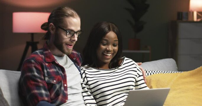 Close up portrait of happy joyful mixed-races young cute couple man and woman chatting and laughing while sitting on sofa in house surfing internet browsing on laptop computer, love concept