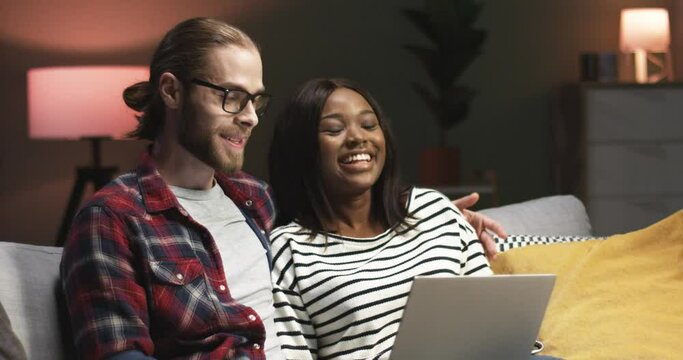 Close up portrait of cheerful young couple of Caucasian man and African American woman speaking and laughing sitting on sofa in house surfing internet, videochatting on laptop computer, love concept