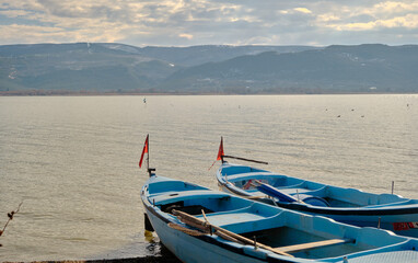 Naklejka premium Small and blue boats near the coast of Lake of Uluabat in Turkey. Small Turkish flag on the boat with lake and mountain background. Bursa. Golyazi (Apollyon). Bursa, Turkey. 22.01.2021.