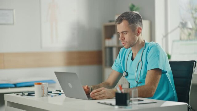 Experienced Male Nurse Wearing Blue Uniform Answering Video Call on Laptop at Doctor's Office