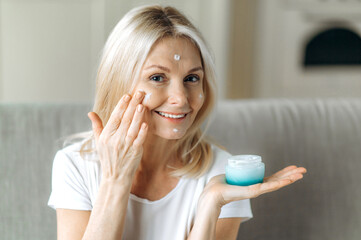 Smiling blonde senior woman apply anti-aging cream to her face. Happy mature woman using cosmetic...